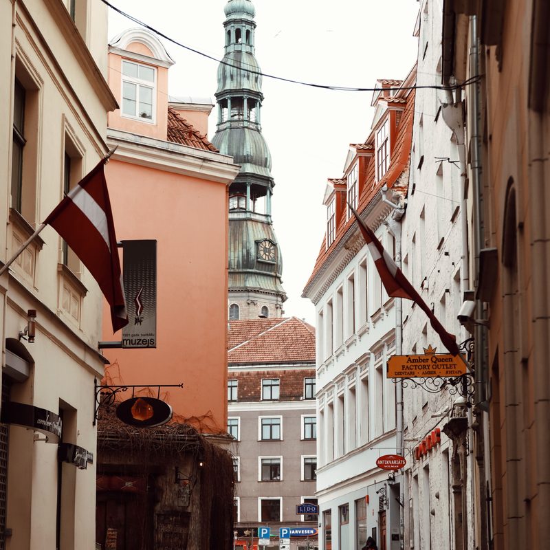 A cobbled street in Riga Old Town with a Latvian flag and St Peter's Church spire