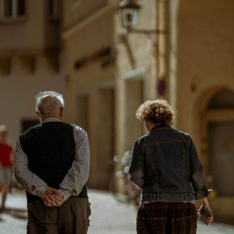 An older couple walking away from the camera along a sunlit cobbled street